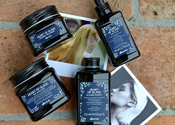 Four black containers of Davines Heart of Glass hair care products, including shampoo and a treatment, arranged on a rustic surface with photographs of blonde hair nearby. Suitable for hair maintenance.