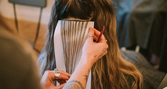 A person with long blonde hair is sitting with their back to the camera, their hair styled with sections folded and pinned using white pieces of paper, resembling a hair treatment technique.