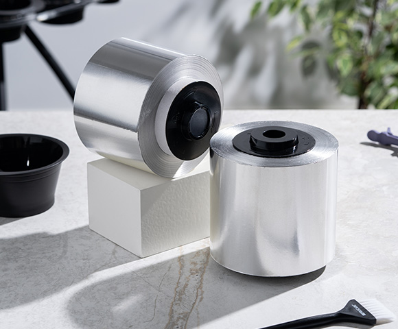 Two rolls of shiny metallic foil positioned on a white block, with a black bowl and a brush nearby on a light-textured surface, showcasing the innovative packaging materials.