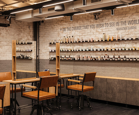 A modern interior of a beauty salon featuring wooden shelves lined with various skincare and beauty products, brown chairs, and a motivational quote by Mahatma Gandhi on the wall.